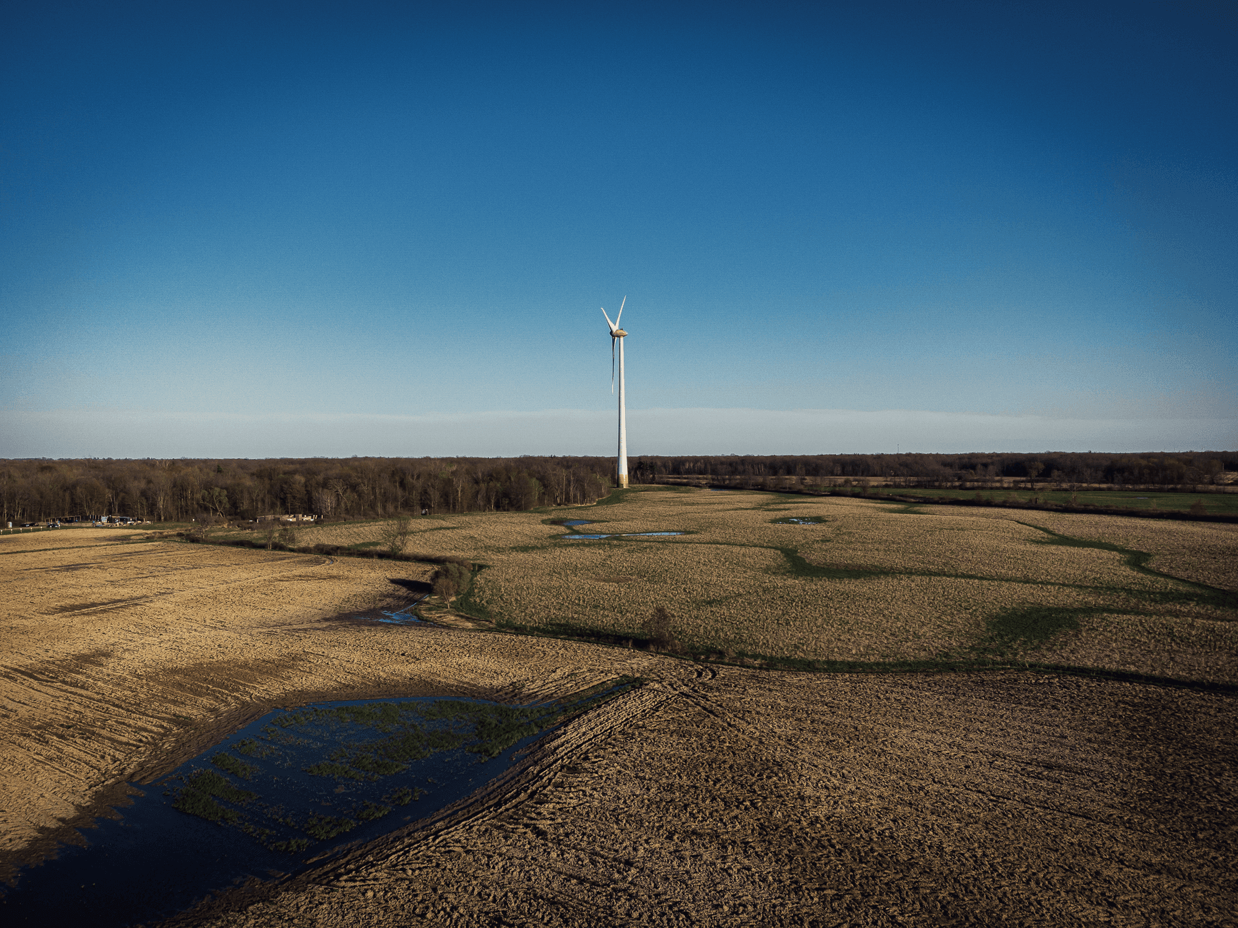 Eén windturbine in een vlak landschap met akkers, plassen en een bomenrij onder een blauwe lucht