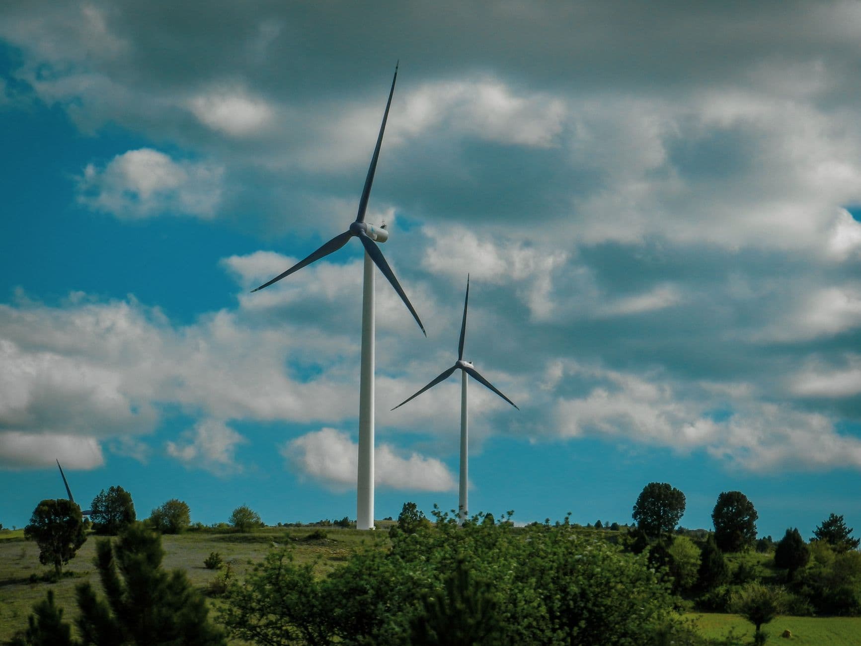 Twee windturbines boven groene bomen onder een blauwe lucht met witte wolken
