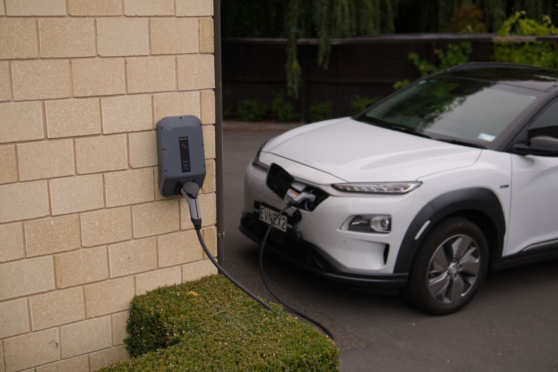 Witte elektrische SUV laadt op via een wandlaadpaal aan een stenen muur op de oprit